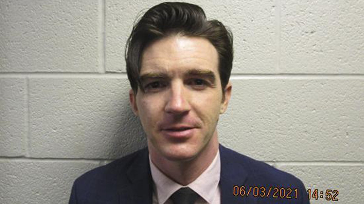 Drake Bell in a suit and tie in front of a white wall mugshot photo 