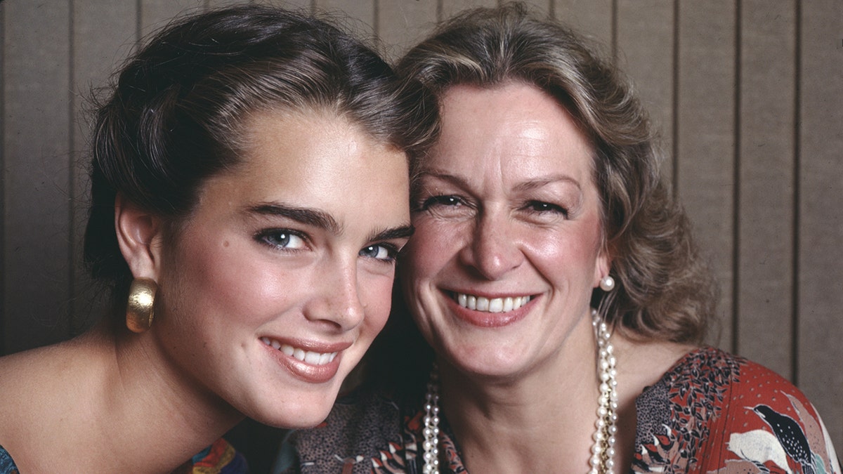 Brooke Shields in a colorful top and large gold earrings smiles next to her mother Teri Shields wearing a pearl necklace and earrings in a photo from 1981