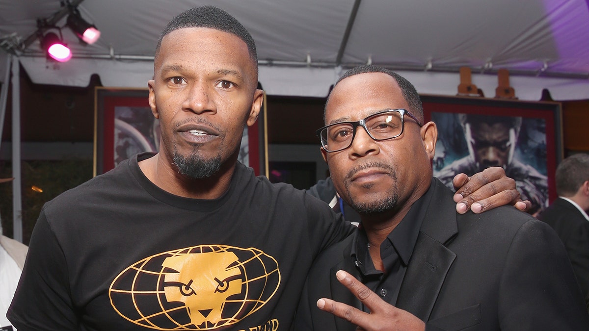Jamie Foxx in a black shirt with a yellow graphic poses for a picture with Martin Lawrence, also wearing a black shirt doing a peace sign
