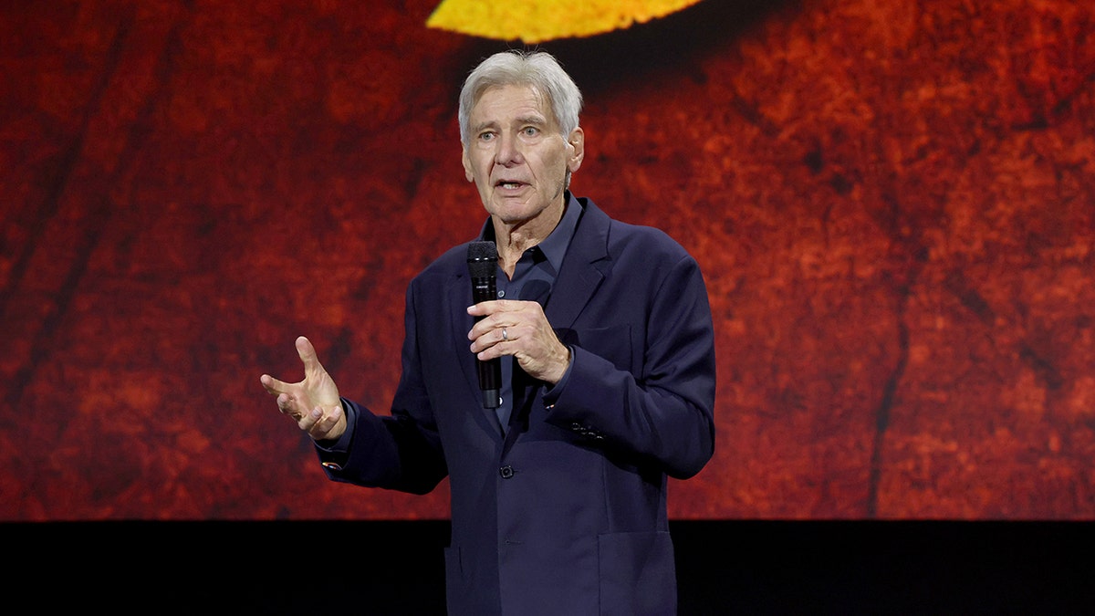 Harrison Ford wearing a dark blue shirt and blazer speaks into a microphone and holds his right hand out while on stage