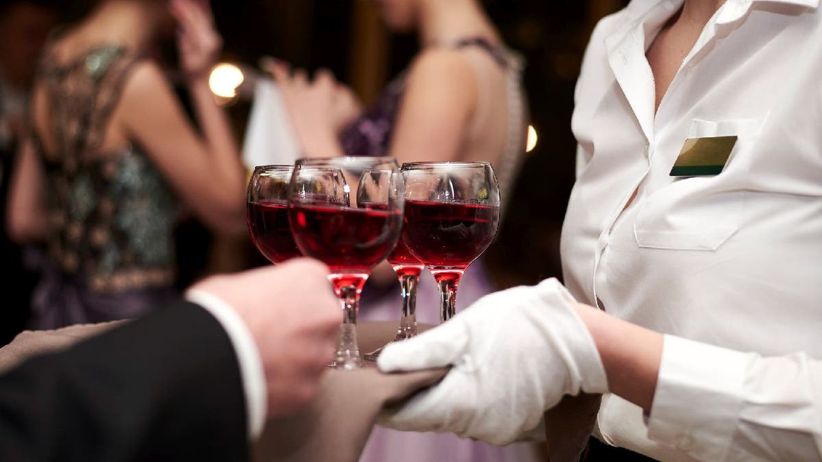 Close-up picture of waiter's hands wearing white gloves holding a tray with red wine, serving alcohol drinks. Catering service at special occasion, event. Hospitality industry concept.