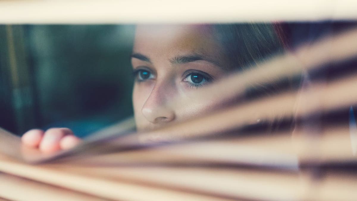 Woman looks out window through blinds