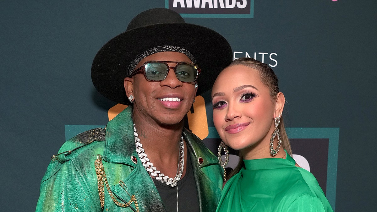 Jimmie Allen in a green jacket and chain necklace and black hat poses next to wife Alexis in a green dress on the carpet