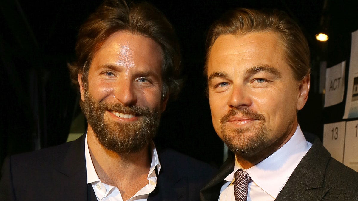 Bradley Cooper with a scruffy beard in a jacket and white shirt smiles next to Leonardo DiCaprio in a suit and tie and white shirt