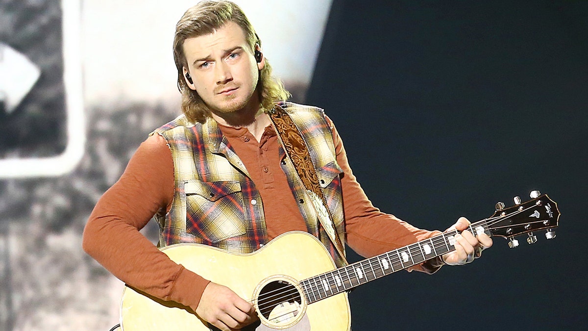 Morgan Wallen strums a guitar on stage while wearing plaid vest and jeans