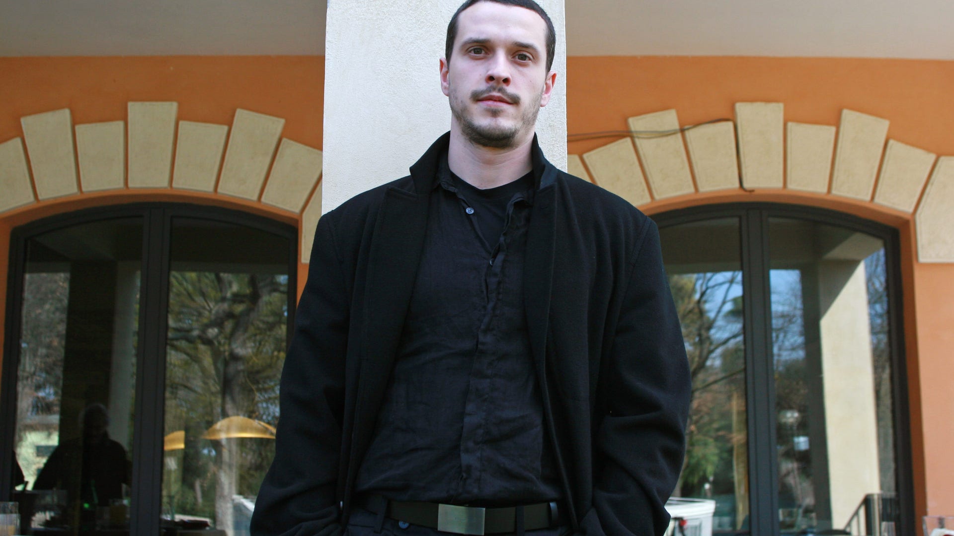 Actor Christo Jivkov wears a black coat and shirt posing in front of a building.