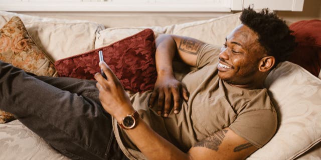 Man laying on his couch while smiling at his phone.