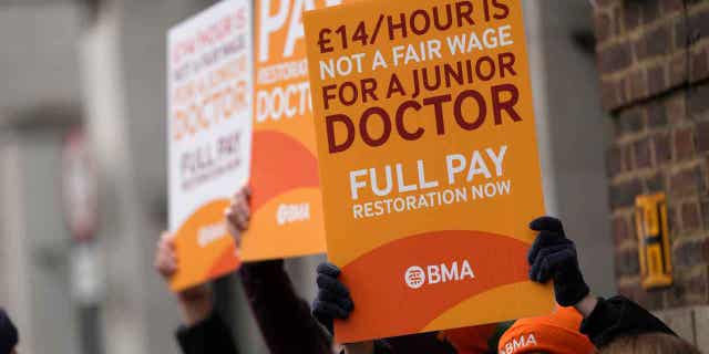 Miles de médicos británicos se preparan para una huelga de 4 días en Gran Bretaña, las autoridades creen que podría ser 'catastrófica' 4 Médicos jóvenes sostienen carteles frente a un hospital en Londres, el 14 de marzo de 2023. Una huelga de cuatro días planeada por médicos en Inglaterra la próxima semana podría provocar el aplazamiento de un cuarto de millón de citas médicas.