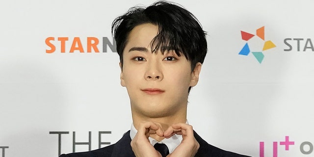 Moon Bin on the red carpet making a heart with both his hands and soft smiling for the camera