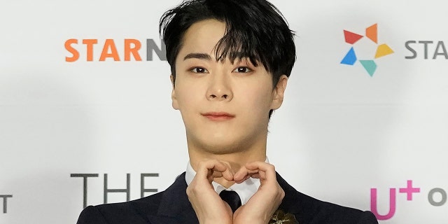 Moon Bin on the red carpet making a heart with both his hands and soft smiling for the camera