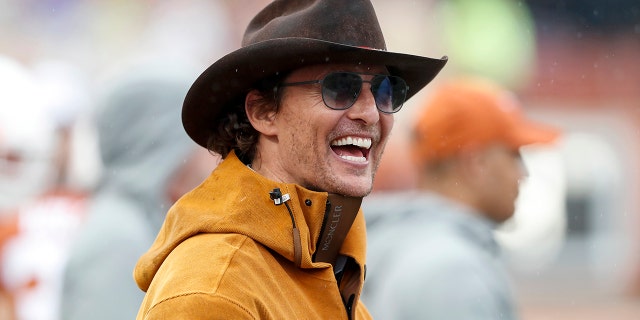 Matthew McConaughey in an orange jacket laughs with a brown cowboy hat on and sunglasses