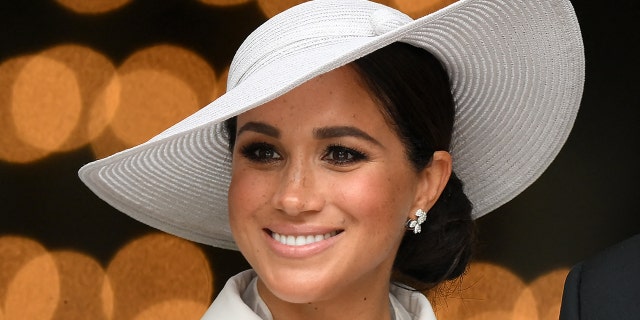 Meghan Markle smiles slightly off-camera in a white hat and matching outfit and earrings