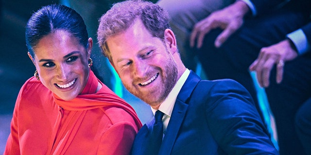 Meghan Markle smiles in a red gown while Prince Harry Leans back towards her and also smiles in a blue suit