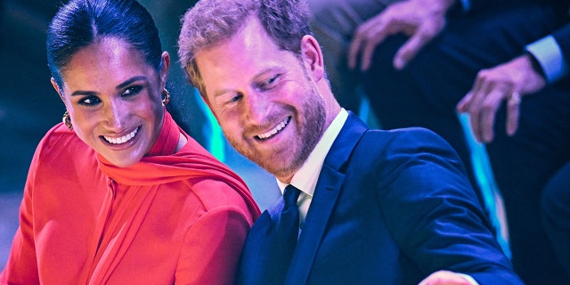 Meghan Markle smiles in a red gown while Prince Harry Leans back towards her and also smiles in a blue suit