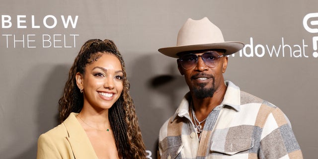 Corinne Foxx in a tan jacket and brown top smiles on the red carpet next to father Jamie Foxx in a patch multi-color shirt with a light tan wide-brimmed hat