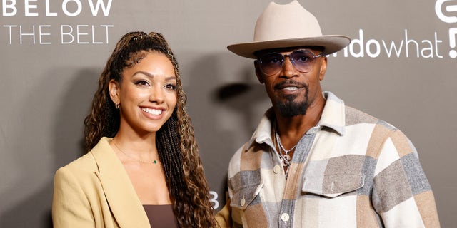 Corinne Foxx in a tan jacket and brown top smiles on the red carpet next to father Jamie Foxx in a patch multi-color shirt with a light tan wide-brimmed hat