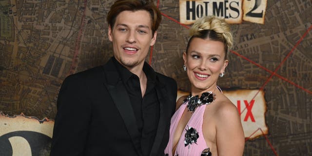 Jake Bongiovi in a black suit poses next to Millie Bobby Brown in a pink dress.