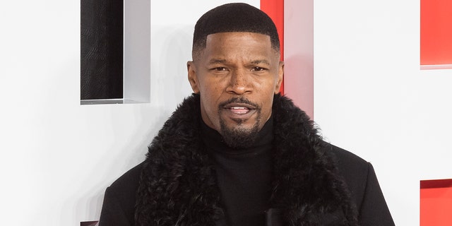 Jamie Foxx in a black turtleneck and coat with a furry trim looks snazzy posing for a photo on the red carpet