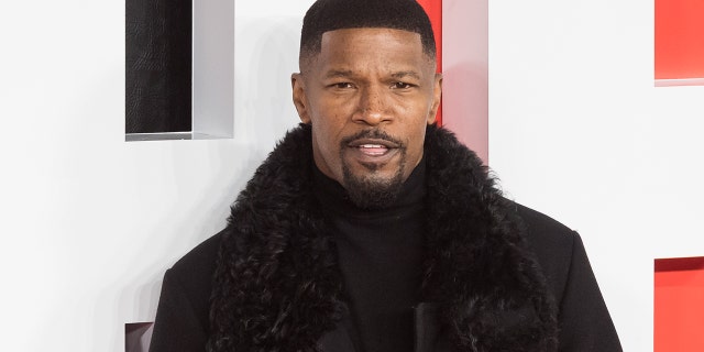 Jamie Foxx in a black turtleneck and coat with a furry trim looks snazzy posing for a photo on the red carpet