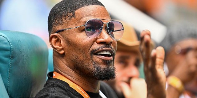 Jamie Foxx raises his hand and waves with an orange lanyard around his neck wearing a black shirt while watching tennis in Miami