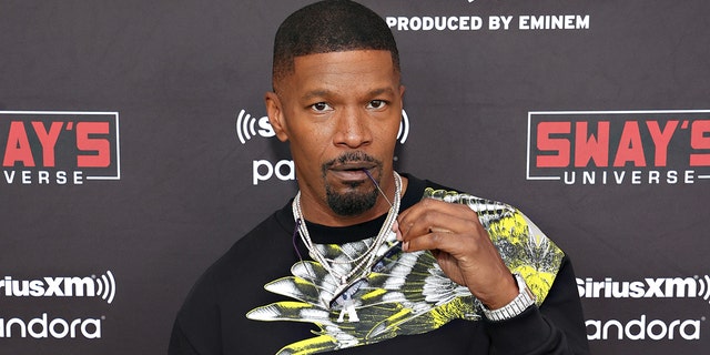 Jamie Foxx bites the end of his glasses on the red carpet with a large chain and black shirt with a white and neon yellow design