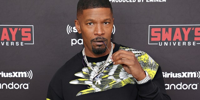 Jamie Foxx bites the end of his glasses on the red carpet with a large chain and black shirt with a white and neon yellow design