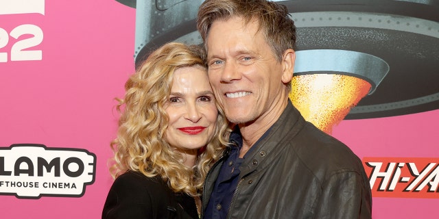 Kyra Sedgwick in a black top and red lipstick cuddles up to husband Kevin Bacon wearing a brown leather jacket and blue shirt flashing a smile