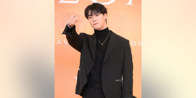 Moon Bin in a black turtleneck and suit and pants with a gold necklace points out in the distance on the carpet