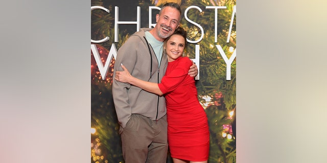 Freddie Prinze Jr. in a grey sweatshirt and light aqua blue shirt is hugged by Rachael Leigh Cook in a red dress on the red carpet for the 