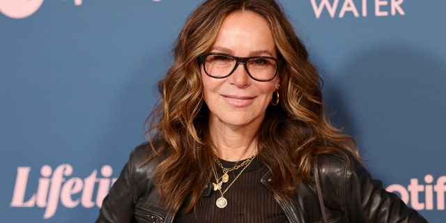 Jennifer Grey in a black leather jacket and black framed glasses smiles on the red carpet