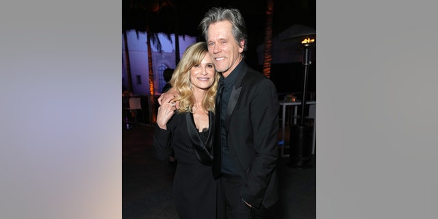 Kyra Sedgwick in a black long jacket holds on to husband Kevin Bacon's hand on her shoulder who is wearing a black suit at the Vanity Fair Oscar party
