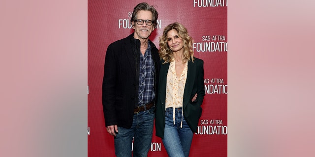 Kevin Bacon in jeans and a black jacket and blue plaid shirt smiles on the red carpet with wife Kyra Sedgwick, also in jeans, a blush blouse and black blazer