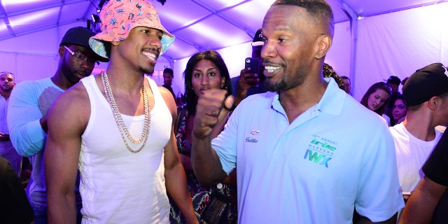 Nick Cannon in a pink hat, a white tank top and large chain smiles at Jamie Foxx who has his fist in the air wearing a blue collared shirt