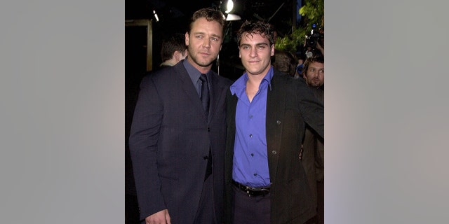 Russell Crowe in a dark suit and tie looks off in the distance to the right posing next to Joaquin Phoenix in a royal blue shirt and dark jacket