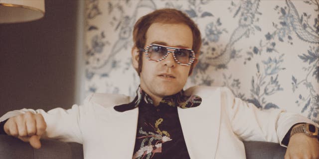 British singer and musician Elton John, wearing a white suit, black shirt with flower motif and multicolored sunglasses, London, November 1973