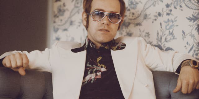 British singer and musician Elton John, wearing a white suit, black shirt with flower motif and multicolored sunglasses, London, November 1973