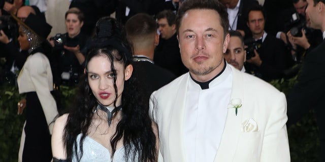 Elon Musk in a white tuxedo and Grimes in a white dress and black accents on the Met Gala red carpet 