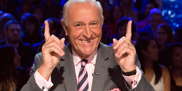 Len Goodman points both fingers up in the air, wearing a grey suit, a pink pocket square, and pink and navy blue striped tie on the set of "Dancing with the Stars"