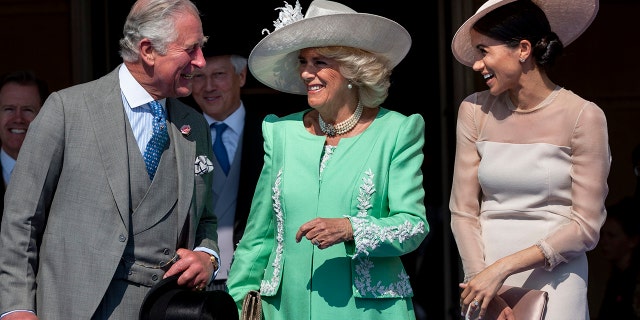 King Charles in a grey multi-piece suit smiles at Meghan Markle in a blush dress and hat, in the middle Camilla smiles in a bright green dress and white hat, looking at Charles