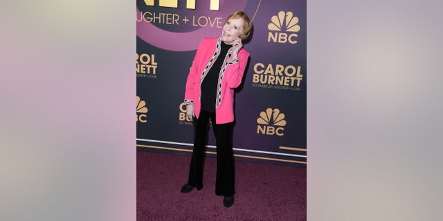 Carol Burnett wearing a black pantsuit with a hot pink blazer holding her ear