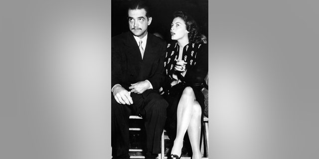 Howard Hughes in a suit sitting next to Ava Gardner who is smoking and talking to him