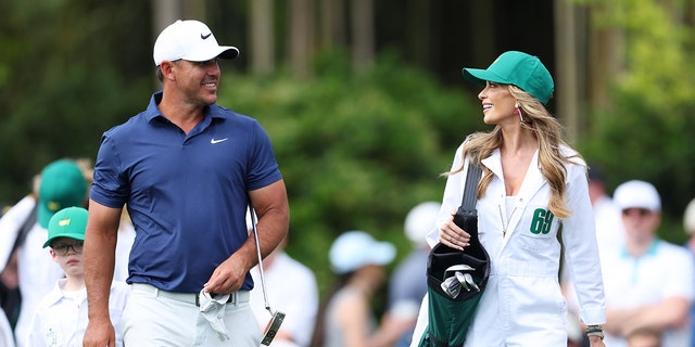 Brooks Koepka in white pants, blue shirt and white cap walking with his wife Jena Sims in a white caddy uniform