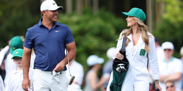 Brooks Koepka wearing white pants and a blue shirt and a white cap walking alongside his wife Jena Sims in a white caddy uniform