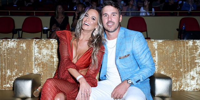 Jena Sims in a low-cut red dress next to Brooks in a white shirt and pants with a bright blue leather jacket