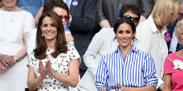 Kate Middleton wearing a white printed dress next to Meghan Markle wearing a blue and white striped blouse holding a wide brimmed hat