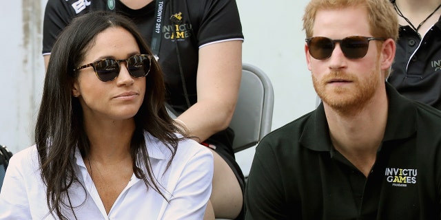 Meghan Markle wearing a white blouse with black sunglasses sitting next to Prince Harry in a black shirt and sunglasses