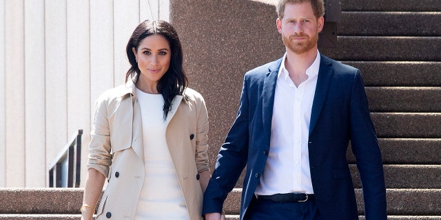 Meghan Markle wearing a white dress with a beige trench coat next to prince harry in a navy blazer and white shirt