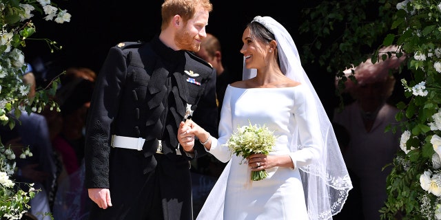 Meghan Markle wearing a white bridal dress holding Prince Harrys hand, who is wearing a suit