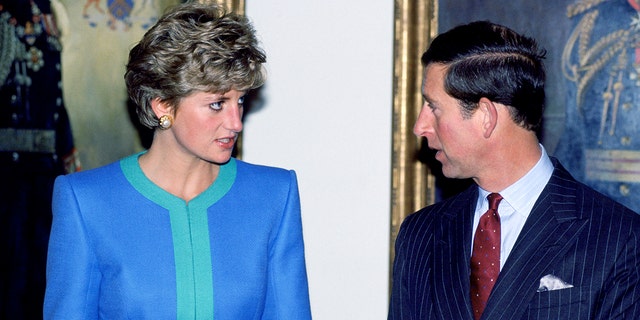 Princess Diana in a blue and green dress looking seriously at King Charles in a suit and red tie