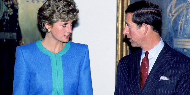 Princess Diana in a blue and green dress looking seriously at King Charles in a suit and red tie